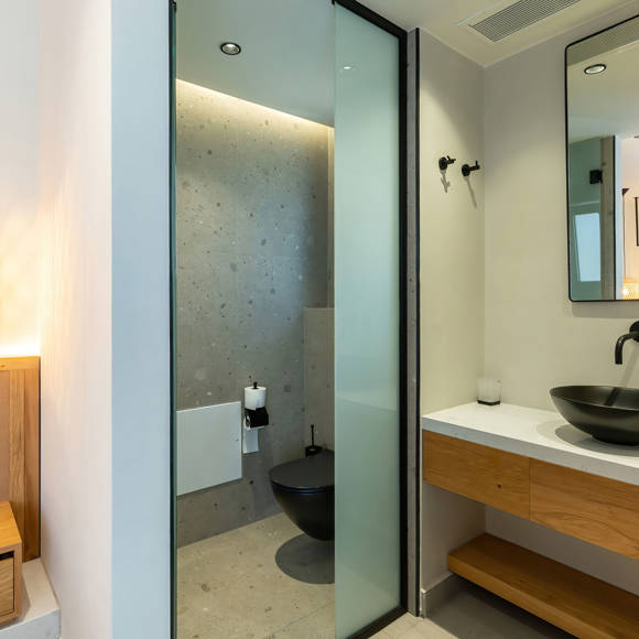 Open bathroom area with a black sink and a big mirror and a separated toilet behind a glass door
