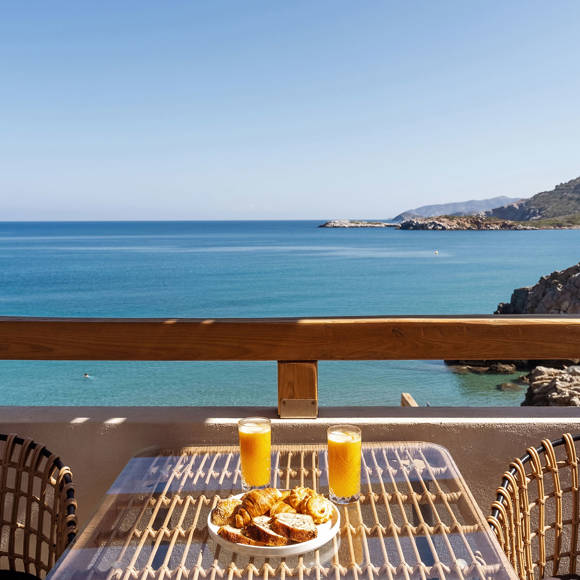 Balcony with a pair of chairs, a table with juice and pastries and a direct sea view with rocky coastline