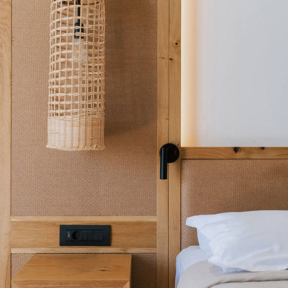 Close-up of a wooden bedside table, a wicker pendant lamp and part of a double bed