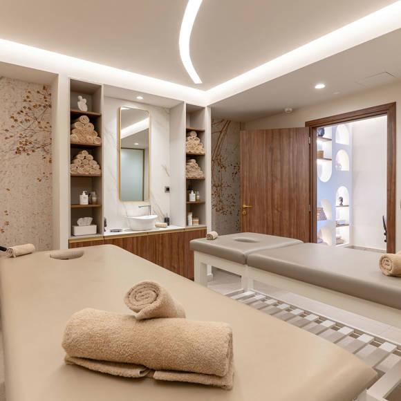 Elegant spa treatment room with two massage beds, soft lighting and built-in shelving with towels