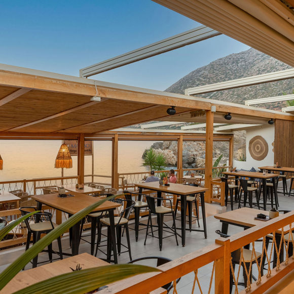 Upper terrace of Mambo Beach Restaurant with wooden roof, high tables and sunset-lit coastal view