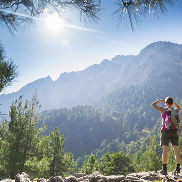 A woman stopping her hike on a sunny day to admire the lush green landscape and distant mountains in Crete