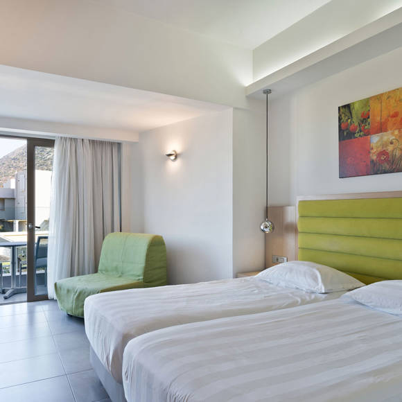 room with two single beds placed side by side with green headboard, a green armchair and a balcony with a view