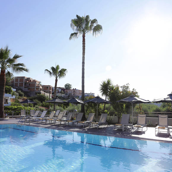 Sunlit swimming pool area lined with loungers and palm trees, offering a relaxing view of the hillside