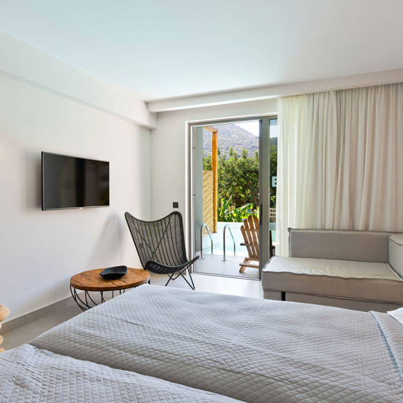Junior suite with two single beds placed side by side, wall-mounted TV, sitting area and access to a pool with garden view