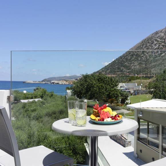 balcony with glass railing, a pair of chairs and a table with drinks and fruit, overlooking the sea and hills