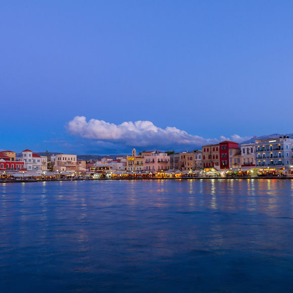 Sunset view of the Chania coastline, seen from the sea, with the city lights reflecting on the water