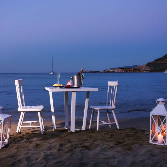 Romantic beach dinner setup at dusk with candle lanterns, a table for two and sea view with anchored sailboats